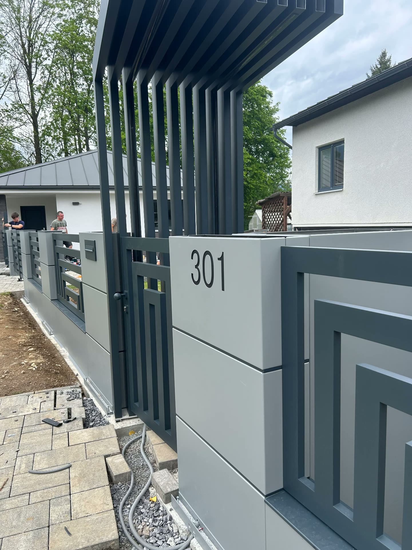 Vstupní branka s lamelovou pergolou, světle šedým modulem a domovním číslem 301
